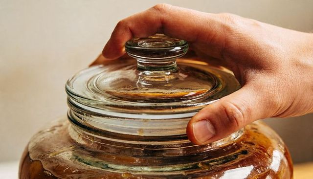 Right hand gripping glass jar lid over amber liquid closeup, kitchen preserving fermentation