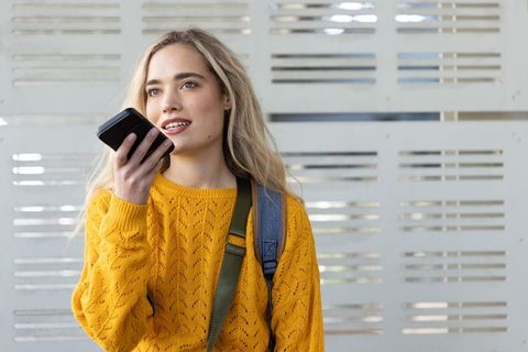 Young woman speaking into smartphone while wearing mustard sweater and backpack