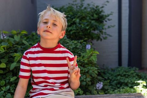 Blond boy sitting on bench showing peace sign in red striped shirt garden portrait