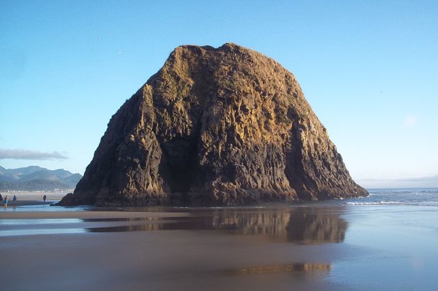 The image showcases Haystack Rock located at Cannon Beach in Oregon, with a clear blue sky in the background. The rock formation is reflected on the wet sand during low tide, highlighting its geological detail and beauty. It is ideal for use in travel, tourism, and natural landscape themes. The prominent landmark in the serene beach environment portrays tranquility and may be used to promote outdoor activities, vacations, and scenic destinations.