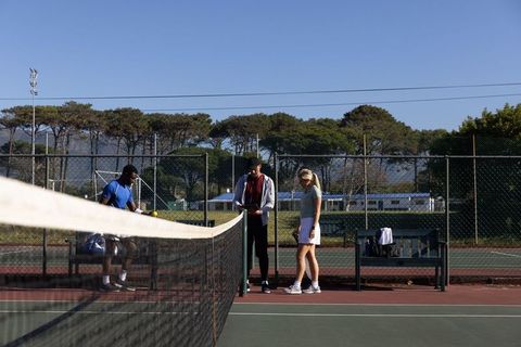 Tennis coach adjusting net strap while players preparing for practice on outdoor hardcourt