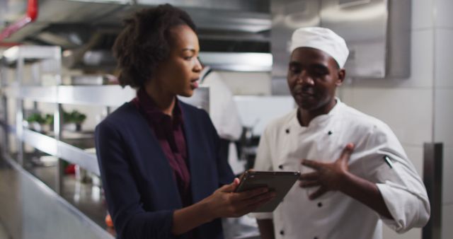 Manager consulting with a chef in a professional kitchen, both engaged in discussion while reviewing information on a tablet. Useful for illustrating workplace collaboration, culinary teamwork, restaurant management, and professional kitchen environments.