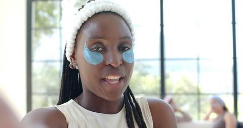 Woman wearing blue hydrogel under-eye patches and terry headband speaking at skincare spa