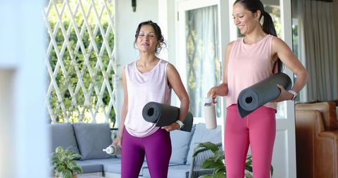 Two women carrying yoga mats in bright home living room wearing pink activewear smiling