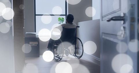 Senior man sitting in wheelchair looking out window in quiet corridor with soft bokeh
