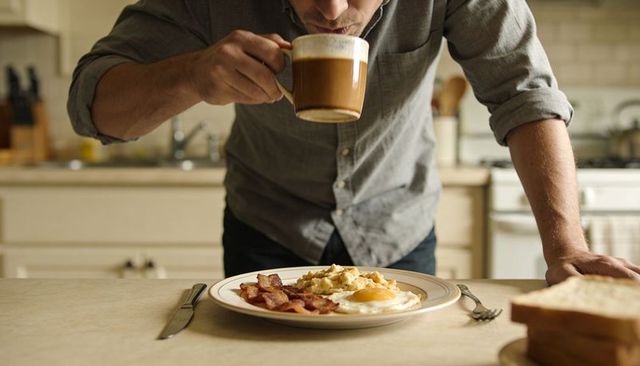 Leaning man sipping coffee over plate of bacon and eggs in cozy morning kitchen