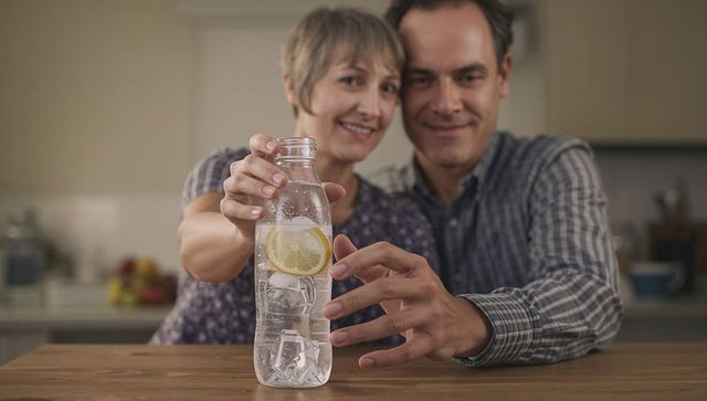 Middle-aged couple sharing lemon water in kitchen leaning together over wooden counter