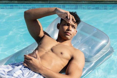 Sunlit african american man relaxing on clear pool float holding drink and shading eyes
