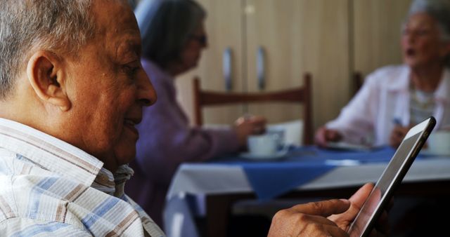 Senior man is seen using a tablet in a retirement home, depicting the integration of modern technology in elderly care. Other seniors are present in the background, suggesting a communal setting. Suitable for illustrating concepts of digital literacy among the elderly, retirement community activities, and modern communication tools for seniors.