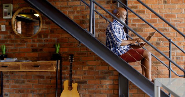African American man sitting on stairs indoors, with copy space. He enjoys a quiet moment at home with a book and a guitar nearby.