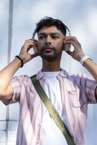 Asian man adjusting over-ear headphones outdoors wearing striped shirt and crossbody bag