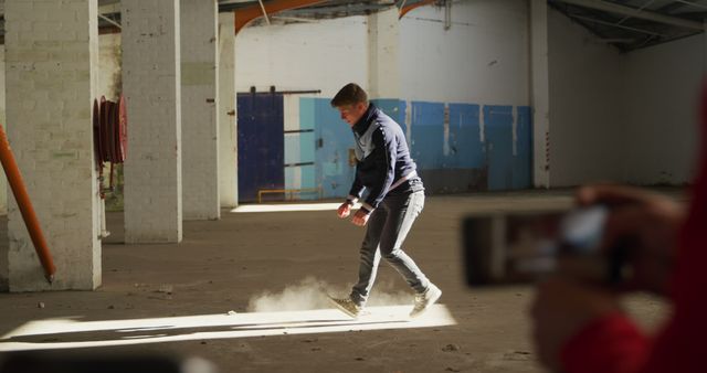 Man wearing casual clothes walks inside an empty, deserted building during daylight. Dust rises from the floor as he steps, adding to the sense of abandonment and adventure. A person nearby captures the moment on a smartphone. Ideal for concepts like urban exploration, adventure, photography tips, and discovering forgotten places.