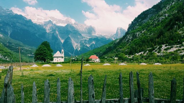 Idyllic summer landscape showing a small village with traditional houses and a church nestled among mountains. The scene includes a lush green field in the foreground and majestic peaks in the distance under a partly cloudy sky. Ideal for use in travel brochures, nature blogs, wallpapers, or publications focusing on rural and serene destinations.