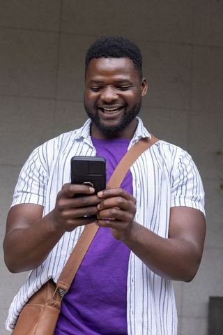 Smiling black man texting on smartphone with crossbody bag, casual summer outfit