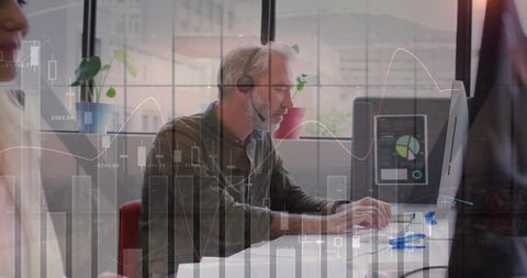 Mature man typing at desktop with headset and financial analytics overlay in modern office