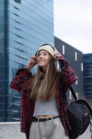 Young woman adjusting headphones on urban rooftop with backpack and cream beanie, city skyline