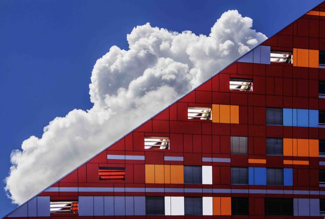 Vibrant modern building exterior set against a blue sky with fluffy clouds. The architectural design features striking geometric patterns in red, blue, and orange tones. Ideal for use in projects related to architecture, urban development, real estate, or contemporary design themes.
