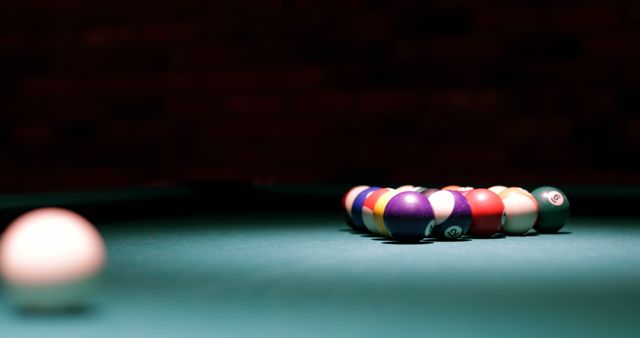 Close-up of a set of billiard balls in a triangle rack on a green billiard table, with the cue ball positioned in the foreground. Perfect for use in articles or advertisements related to indoor sports, leisure activities, bar games, and recreational pursuits. This visual can be used to promote pool halls, game rooms, or products related to billiards.