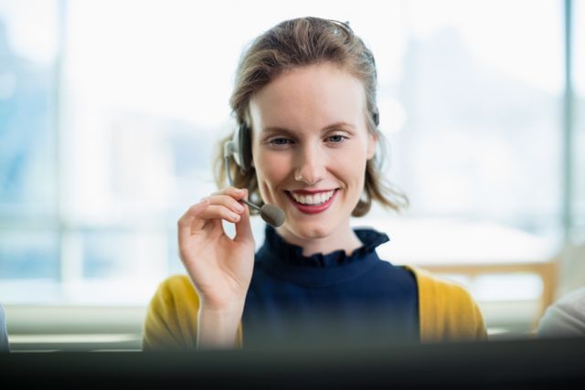 Young woman wearing headset, smiling while working in a call center. Ideal for illustrating customer support, telemarketing services, business communication, and professional work environments.