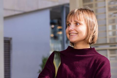 Young woman standing near glass entrance smiling looking sideways in burgundy sweater