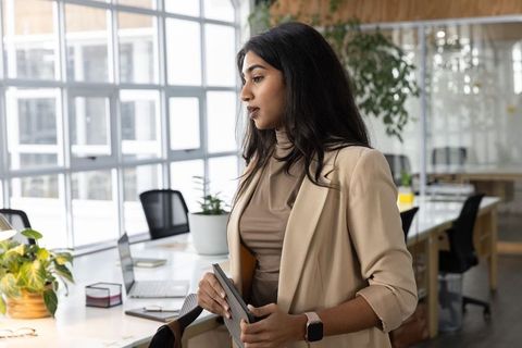 Indian professional standing at open-plan desk holding tablet and gazing left