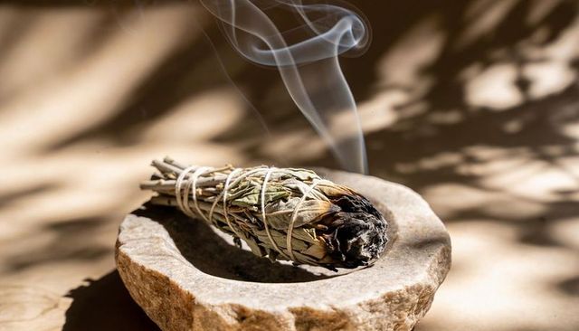 Burning white sage smudge bundle emitting smoke on stone dish with dappled shadows