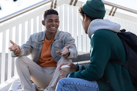 College friends chatting on outdoor steps holding coffee and laughing during campus break