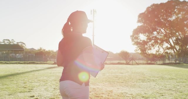 Image shows a female coach standing with a clipboard in hand on a sports field at dusk. The backlighting creates a silhouette effect with the warm light of the setting sun. Ideal for use in articles or advertisements relating to sports coaching, female empowerment in sports, outdoor training, or team leadership.
