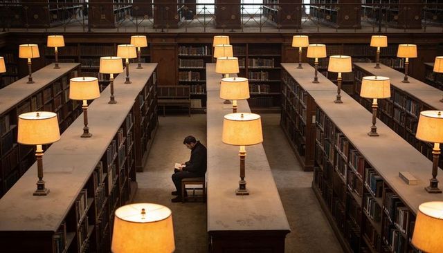 Man Reading Alone in Historic Library Under Rows of Warm Desk Lamps and Mezzanine