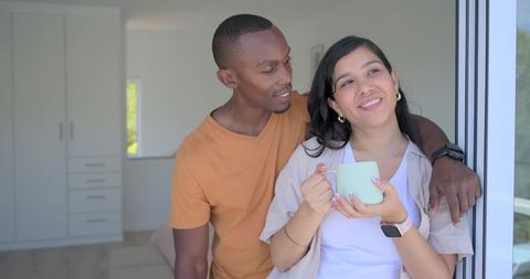 Leaning couple smiling and holding mug by sliding glass door in modern apartment with smartwatches