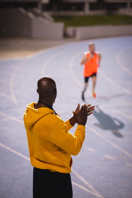Back view of an African American male coach clapping hands while encouraging a professional runner training on a sports stadium track. Ideal for use in articles or advertisements related to sports coaching, athletic training, motivation, teamwork, and fitness programs.
