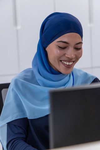 Smiling african american professional wearing blue hijab working on laptop in modern office