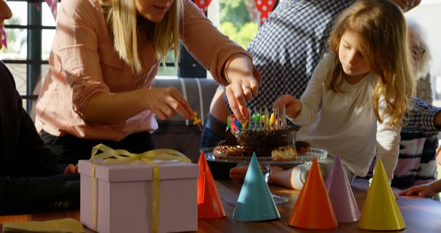 Family members are celebrating a child's birthday by lighting candles on a cake. Colorful party hats and a wrapped gift are on the table, capturing a festive and joyful moment. Ideal for use in birthday, celebration, family bonding, and childhood joy contexts.