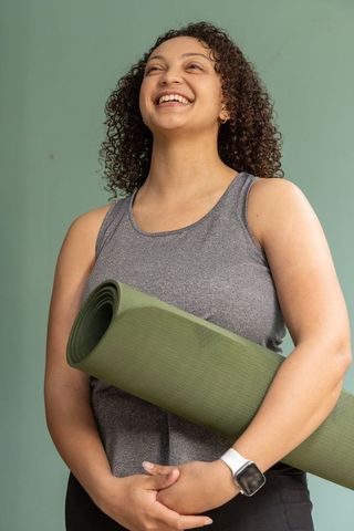 Smiling african american woman holding green yoga mat, wearing smartwatch and tank top