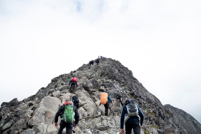 A group of hikers is climbing a steep rocky mountain trail. Each hiker is carrying a backpack as they navigate the rugged terrain, determined to reach the summit. This image can be used for promoting outdoor adventure activities, fitness and health initiatives, or showcasing the beauty of nature and challenge of mountain trekking.
