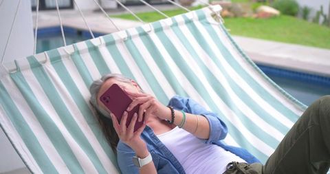 Mature woman relaxing on striped hammock in backyard by pool using smartphone and smartwatch