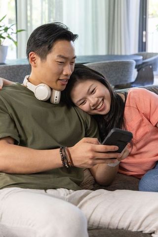 Asian couple cuddling on sofa smiling and viewing smartphone in modern cozy living room