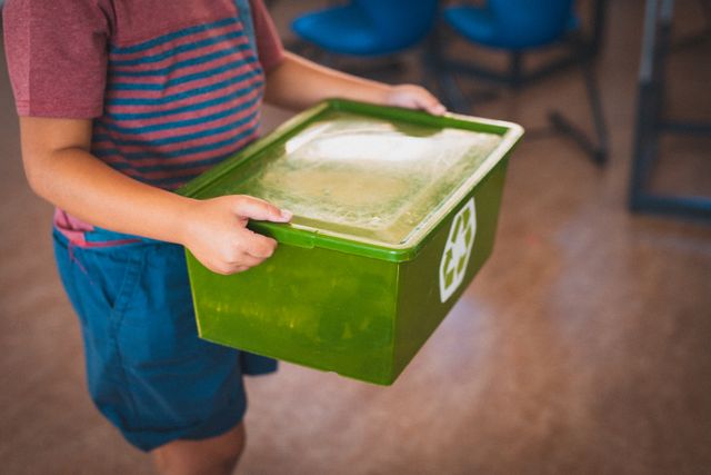 Schoolboy holding green recycling box in classroom. Ideal for educational materials on sustainability, environmental awareness campaigns, and school recycling programs.