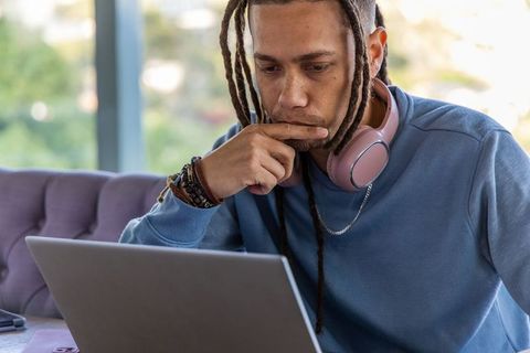 African american man working on laptop wearing pink headphones on purple sofa focused