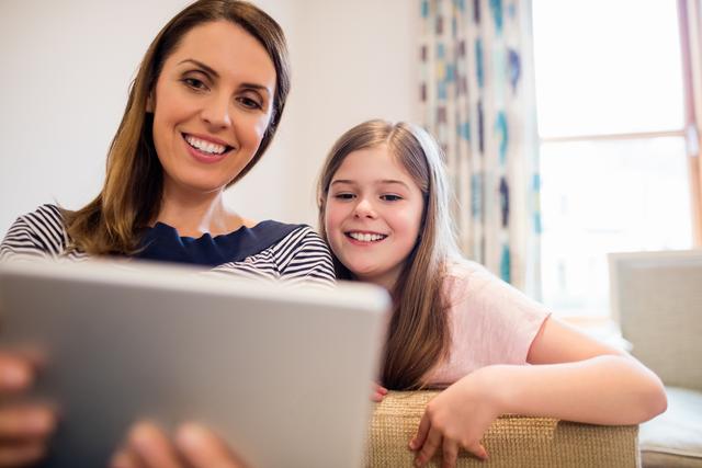 Mother and daughter sitting on sofa using digital tablet in living room at home