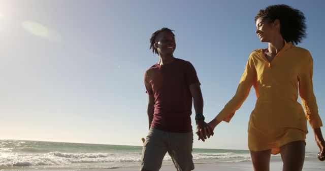 Happy african american couple walking, holding hands and bottles on sunny beach with copy space. Summer, vacations, free time, food and drink, togetherness and lifestyle, unaltered.