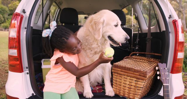 A young girl playing and interacting with a dog in an open car trunk. The setting suggests a picnic with a wicker basket and a guitar present. Ideal for advertisements, blogs on outdoor activities or family time, and social media posts promoting quality time with pets.