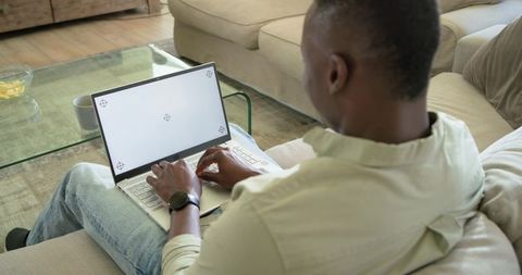 African American man typing on laptop on sofa at home with mug and snacks