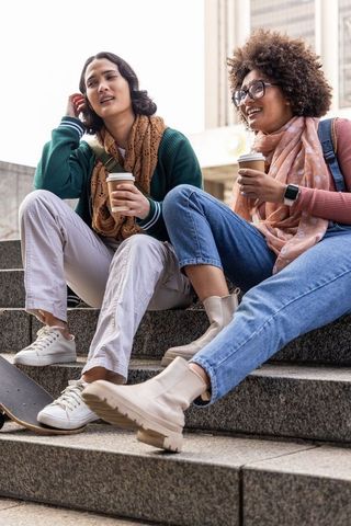 Multicultural young women sipping coffee and chatting on urban steps with skateboard