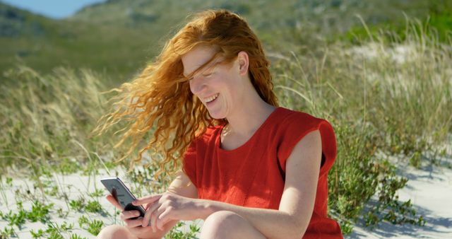 Redheaded woman wearing red shirt, relaxed and smiling while using smartphone at a scenic beach. Ideal for depicting leisure, summer vacations, and casual connection with technology. Suitable for themes of outdoor activities, happiness, digital lifestyle, and enjoying nature.
