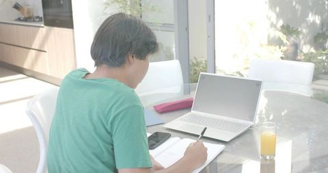 Asian man writing and studying at home dining table with laptop, smartphone and orange juice