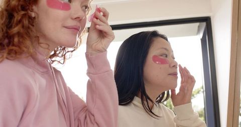 Diverse friends applying pink under-eye patches at vanity mirror for skincare routine