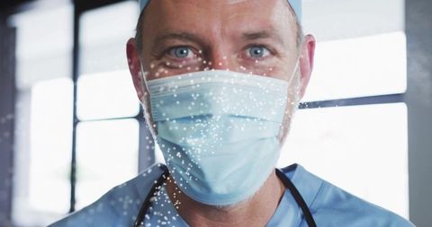 Male clinician wearing surgical mask and scrub cap standing behind droplet-speckled glass