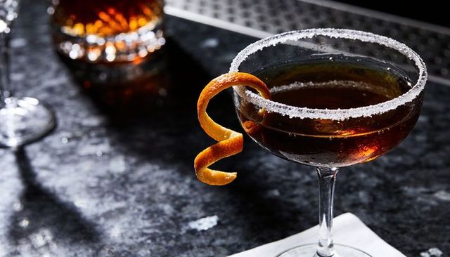 Sugared coupe glass holding dark amber cocktail with citrus twist on glossy stone bar