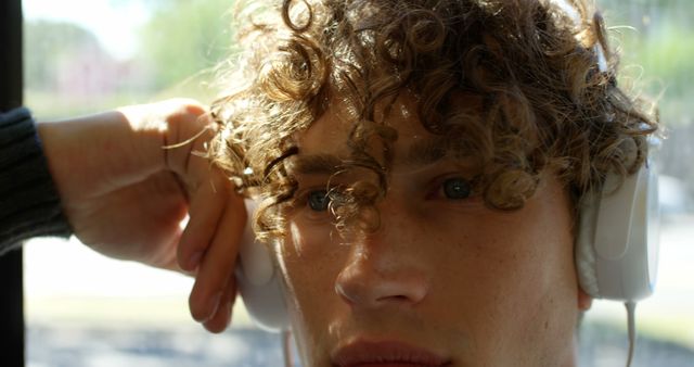 Close-up of a young man with curly hair wearing headphones and looking into the distance, capturing a serene and thoughtful moment. This image can be used in blogs or articles discussing youth, music, personal moments, technology, or modern lifestyle. It is ideal for promoting audio products or creating mood boards that delve into personal experiences or contemporary youth culture.